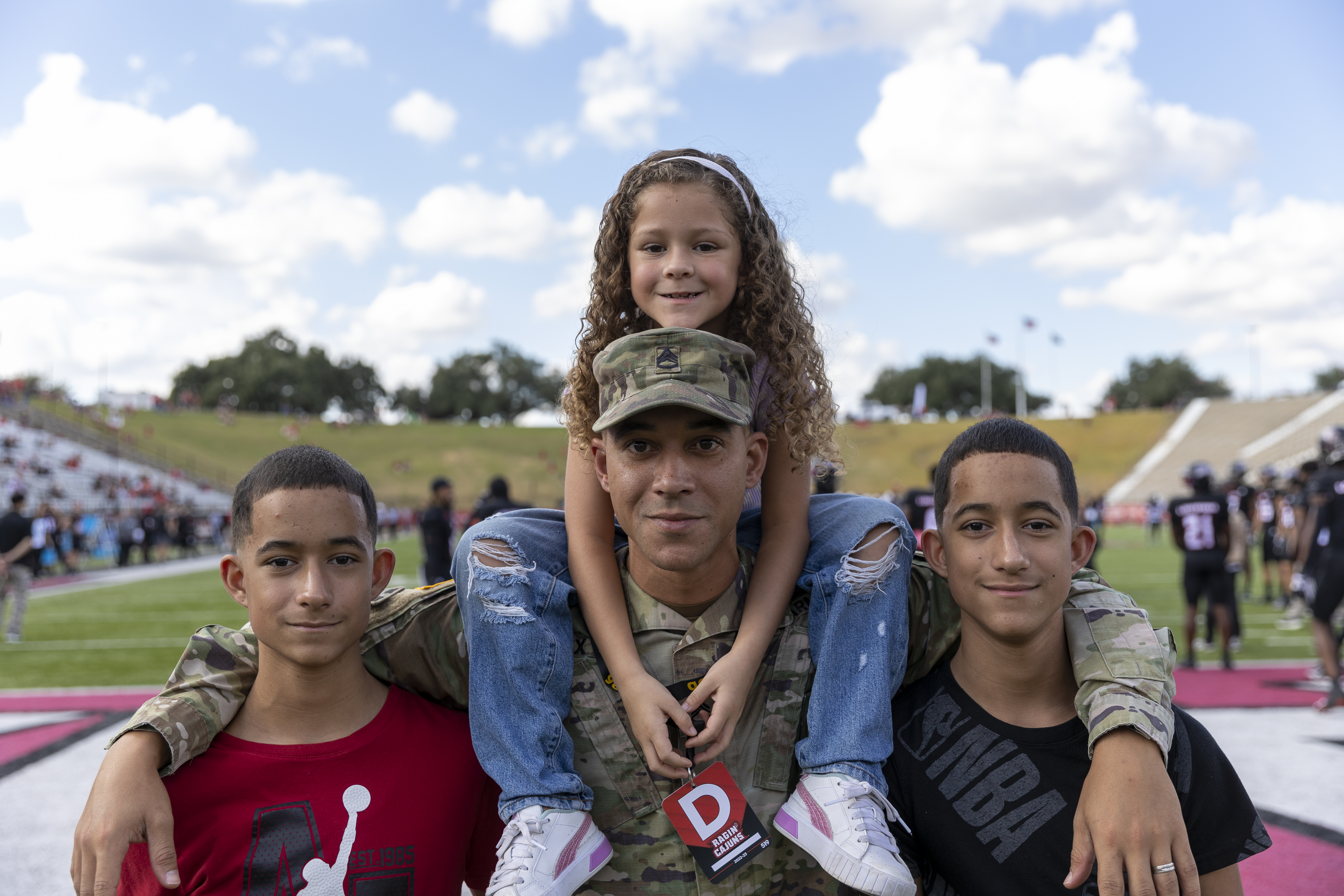 Man in veteran attire has his arms around two boys as a girl sits on his shoulders, a football field with players in the background